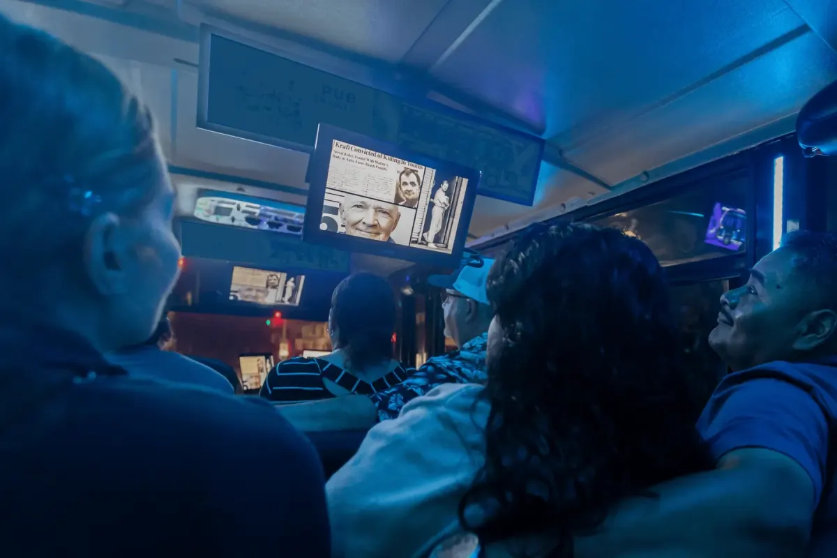 Guests in blue light inside the tour bus watching a screen showing a newspaper clipping